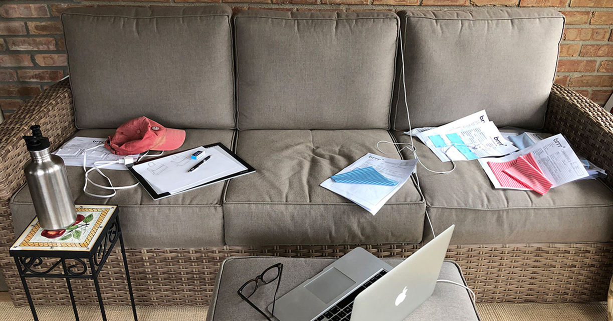 color photograph of laptop computer on small table adjacent to couch strewn with miscellaneous items–loose papers, a hat, mail..