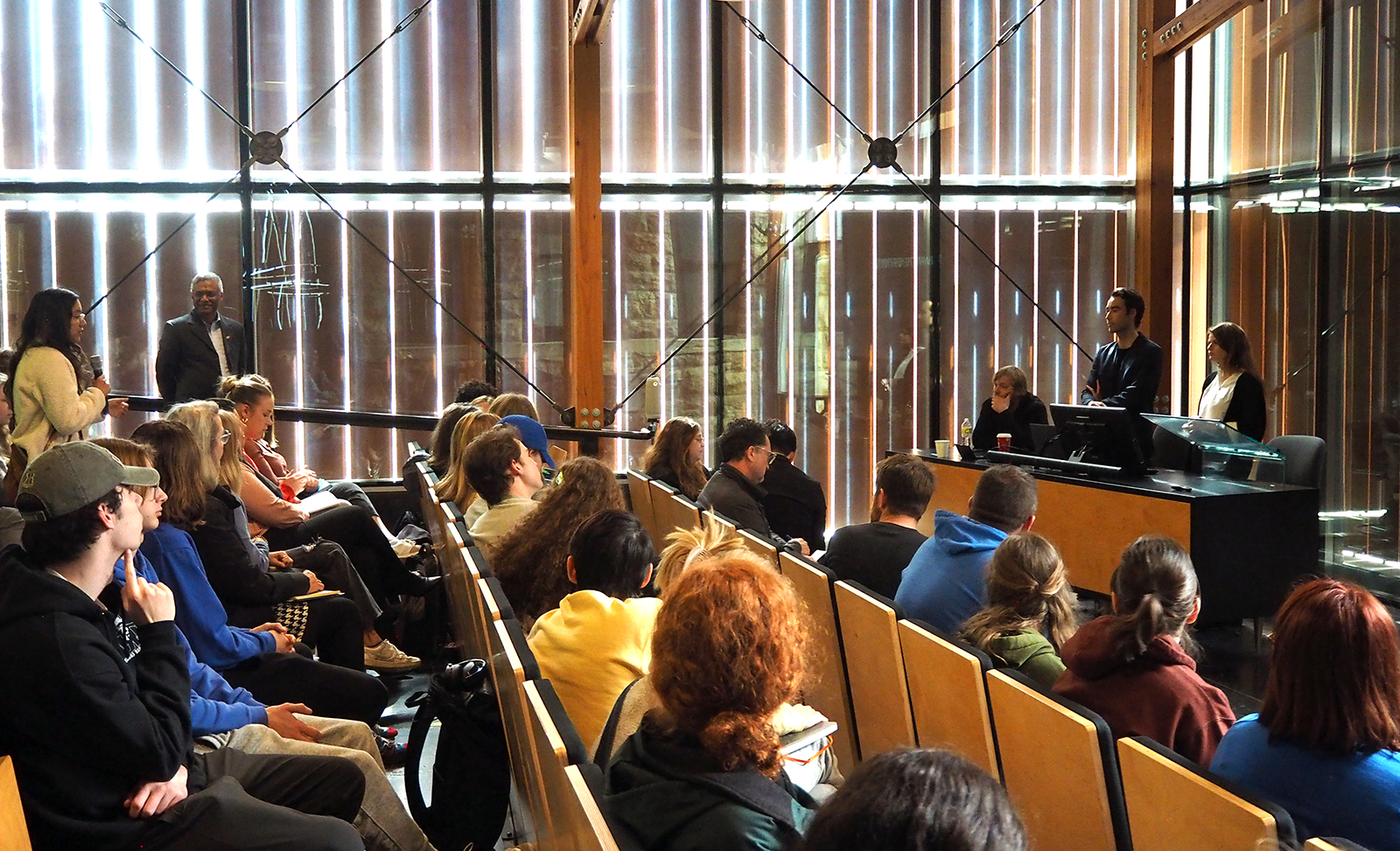 Student holds microphone posing a question to presenters in full, glass-walled auditorium. Sunlight enters the space through vertical wood louvers.