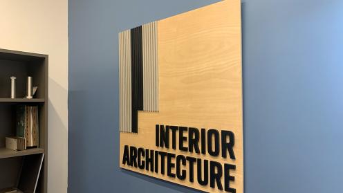 color photograph shows natural wood signage with black lettering that reads 'Interior Architecture' installed on blue wall.